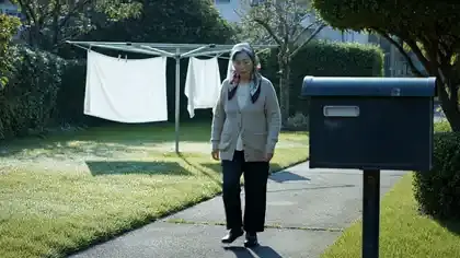 An older woman in a cardigan walking up a garden path toward a mailbox in morning light, seamless loop